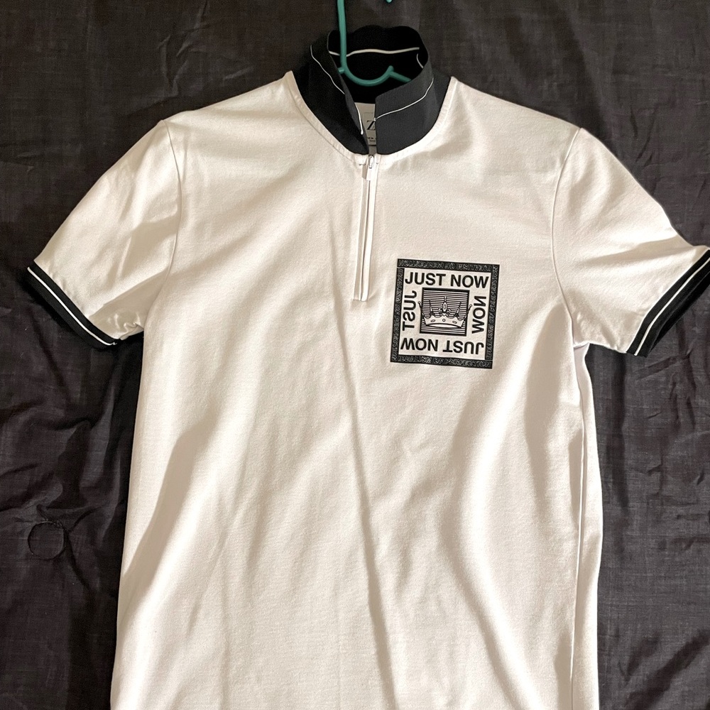 A white and black polo style t-shirt with the logo that says “Just Now”.